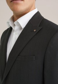 Vīrietis, kurš valkā tumšu svītrainu uzvalka žaketi ar izgriezumu un baltu krekli, tuvplāns uz krūtīm un apakšējo sejas daļu.