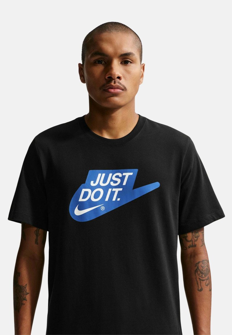 Hombre con cabello corto y bigote, que lleva una camiseta negra con el logo azul de Nike y el texto "JUST DO IT.". Tatuajes visibles en ambos brazos.