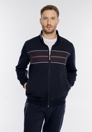 Mężczyzna w granatowej kurtce na zamek z białym i czerwonym paskiem na wysokości klatki piersiowej oraz dopasowanych granatowych spodniach, ręce w kieszeniach, neutralne tło.