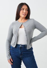 Grijze gebreide cardigan met een ronde halslijn en knoopsluiting, gecombineerd met een witte top en blauwe jeans, met lange mouwen en een getailleerd ontwerp.