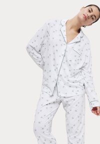 Witte pyjamastel met een patroon van kleine groene stervormige figuren. Het shirt heeft een kraag, een borstzak en contrasterende biezen.