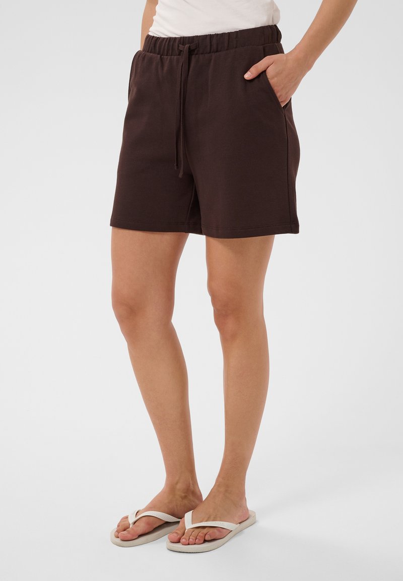 Femme portant un short marron foncé à taille élastique avec des poches à cordon et des tongs blanches, debout sur un fond uni.