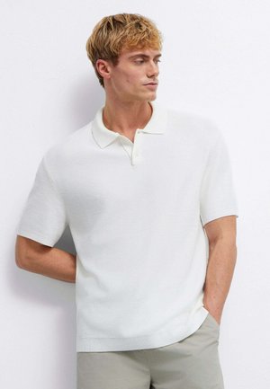 Junger Mann mit blondem Haar, der ein weißes kurzärmeliges Poloshirt und helle Hosen trägt, steht vor einer weißen Wand.