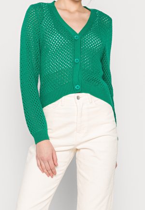 Femme portant un cardigan vert en maille ajourée boutonné et un pantalon blanc taille haute, debout avec une main reposant sur sa jambe.