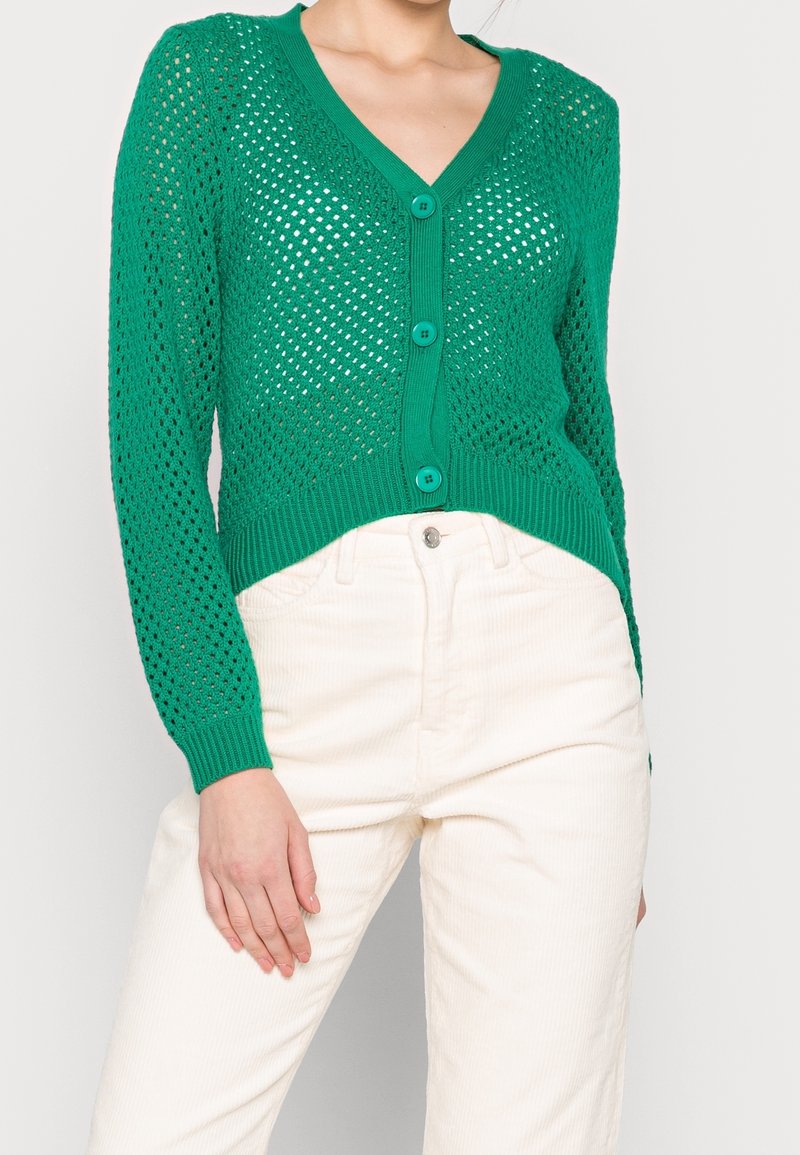 Femme portant un cardigan vert en maille ajourée boutonné et un pantalon blanc taille haute, debout avec une main reposant sur sa jambe.