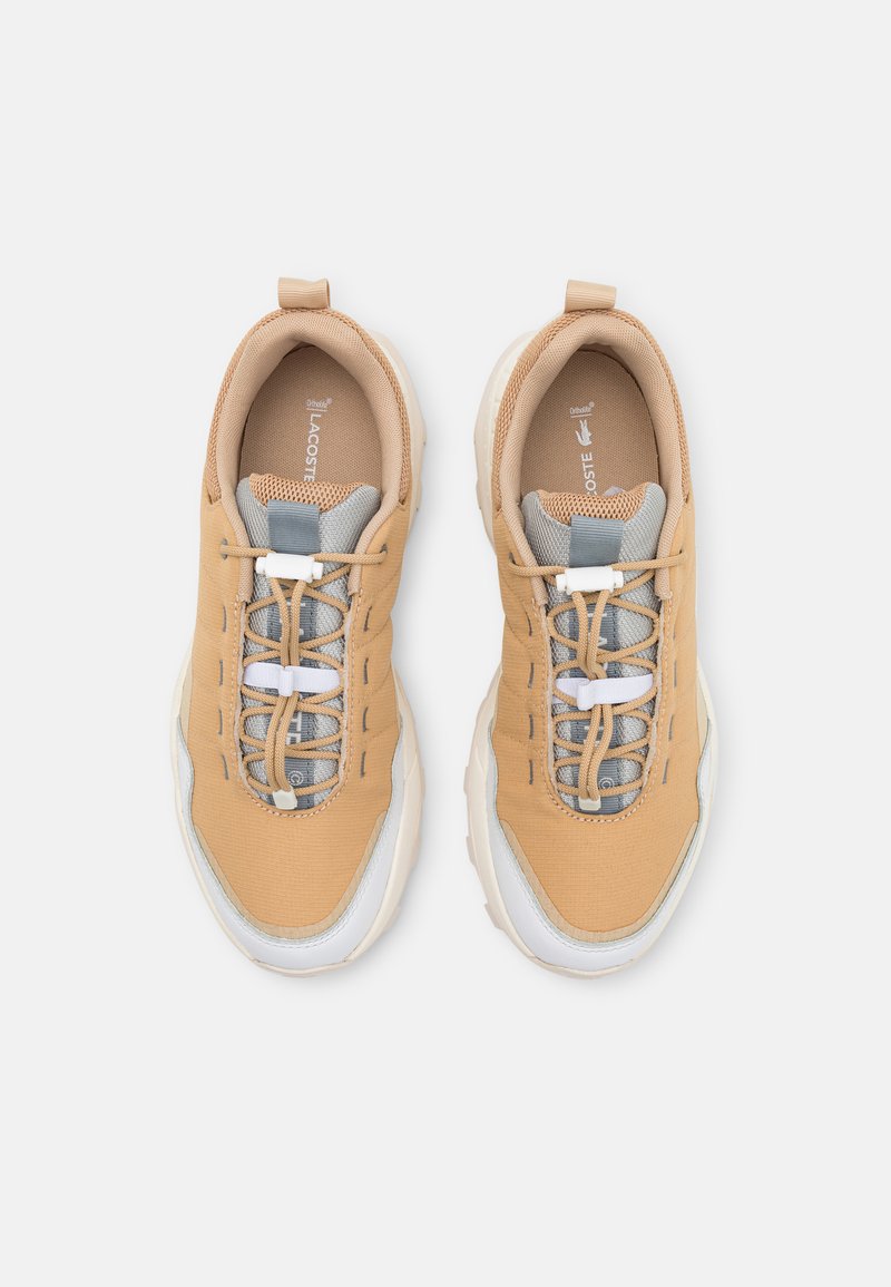Des baskets beiges présentent une tige en maille et en textile, des accents gris, des lacets ronds et une semelle blanche avec des détails texturés.