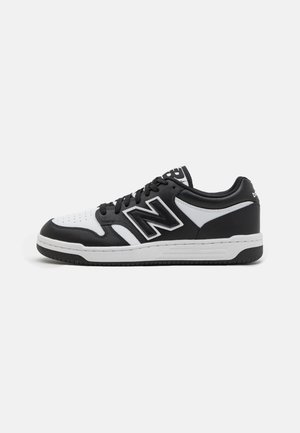 Baskets basses New Balance noires et blanches avec grand logo latéral, lacets noirs et semelle blanche à crampons noirs.