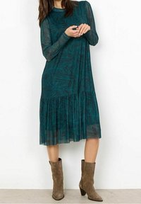 Vestido de manga larga en color teal con un patrón de hojas y falda de volantes. Lleva unas botas de tobillo marrones, tiene un escote redondo y mangas transparentes.