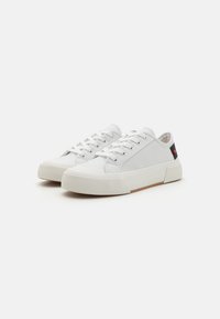 Sneaker bianche in canvas con punta rotonda, suola piatta in gomma e design con lacci. Presentano una parte superiore texturizzata e un piccolo logo rosso sul tallone.