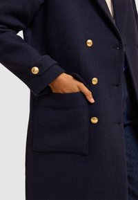 Cappotto navy in lana con design doppiopetto, bottoni dorati e tasca frontale con finitura testurizzata.