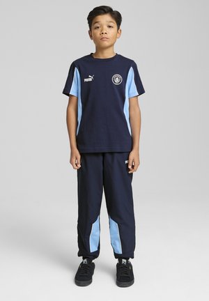 Marineblauwe sporttrackuit met een T-shirt met korte mouwen en lichtblauwe accenten, en bijpassende broek met blauwe zijpanelen. Zwarte sneakers.