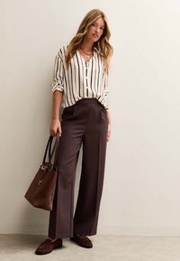 Gestreepte crèmekleurige en zwarte overhemd met knopen, bruine wijde broek, bruine suède schoudertas en bijpassende loafers in een neutrale setting.