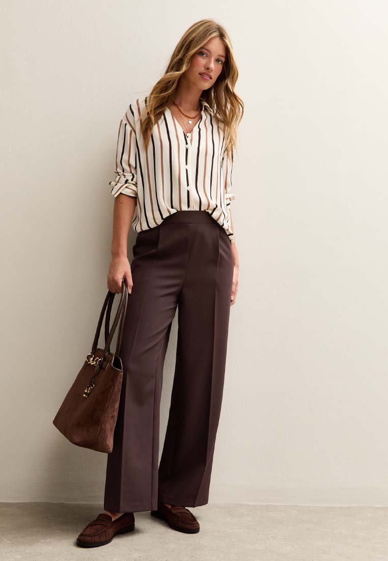 Gestreepte crèmekleurige en zwarte overhemd met knopen, bruine wijde broek, bruine suède schoudertas en bijpassende loafers in een neutrale setting.