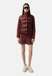 Burgunderfarbene Puffer-Weste mit hohem Kragen und Knopfverschluss, kombiniert mit passenden Shorts und klobigen, cremefarbenen Platform-Loafern.