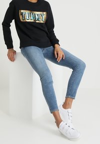 Czarna bluza z kapturem z logo "TOMMY" zrobionym ze złotych i turkusowych cekinów, zestawiona z jasnoniebieskimi obcisłymi jeansami i białymi sneakersami na rzepy.