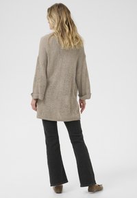 Beige Strickjacke mit lockerer Passform und aufgerollten Ärmeln, kombiniert mit dunklen Schlagjeans und braunen Slippern. Sanfte Textur, lässiges Design.
