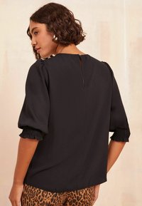 Blusa nera con maniche corte a sbuffo con polsini smockati. La parte posteriore presenta un dettaglio a chiave con una piccola chiusura a bottone. Tessuto di texture liscia.