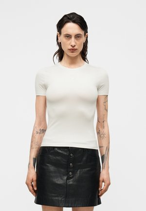 Γυναίκα με σκούρα μαλλιά που φοράει ένα απλό λευκό εφαρμοστό κοντομάνικο μπλουζάκι και μια μαύρη δερμάτινη φούστα με κουμπιά μπροστά, δείχνοντας τατουάζ και στα δύο χέρια.