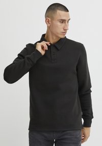 Svart langermet polo-skjorte laget av mykt stoff, med krage og tre knapper, ribbede mansjetter og en avslappet passform.