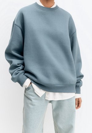 Personne portant un sweat-shirt oversize bleu clair sur une chemise blanche, associé à un jean délavé clair, debout avec une main dans la poche.