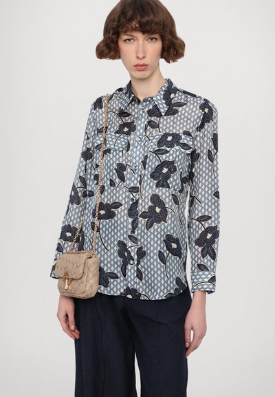 Bluse mit Blumenmuster in Hellblau und Marineblau, ausgestattet mit zwei Vordertaschen und einer lockeren Passform, kombiniert mit einer gewebten beigen Handtasche.