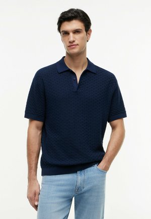 Mand iført navy tekstureret kortærmet polo og lyseblå jeans, stående med den ene hånd i lommen mod en hvid baggrund.