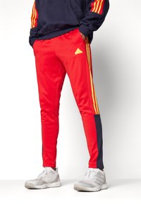 Pantaloni sportivi rossi con strisce gialle e un pannello laterale blu navy, dotati di una cintura elastica e un piccolo logo sulla coscia.