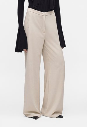 Pantalon beige clair à jambes larges avec poches latérales, porté avec un haut noir à manches longues avec poignets évasés et chaussures noires pointues.