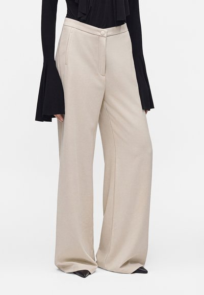 Pantalon beige clair à jambes larges avec poches latérales, porté avec un haut noir à manches longues avec poignets évasés et chaussures noires pointues.