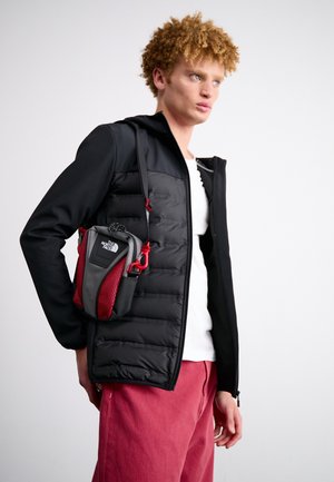 Jeune homme aux cheveux bouclés portant une veste noire, une chemise blanche et un pantalon rouge, portant un petit sac bandoulière North Face noir et rouge.