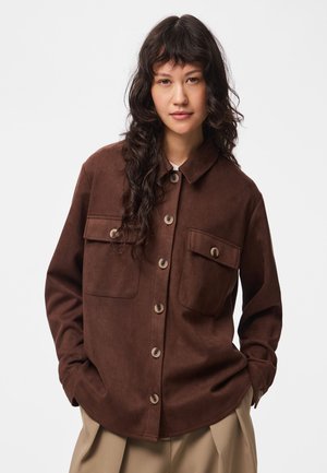 Femme aux longs cheveux bouclés et foncés, portant une veste marron boutonnée avec deux poches poitrine et un pantalon beige, debout devant un fond uni.