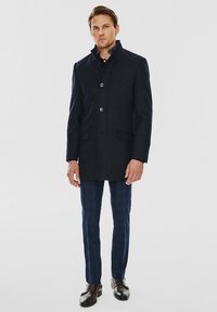 Niebieski płaszcz wykonany z mieszanki wełny, z wysokim kołnierzem, trzema guzikami i bocznymi kieszeniami. W połączeniu z niebieskimi spodniami w kratę i skórzanymi butami.