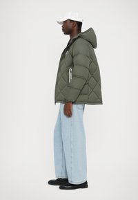 Maslinasto zelena puffer jakna s kapuljačom, dijamantno quilted dizajnom, patentnim zatvaračima i prednjim džepovima. Uparena s svijetloplavim širokim trapericama i crnim cipelama.