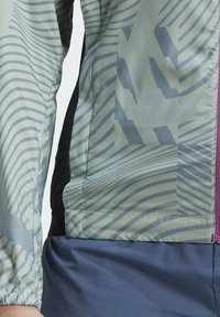 Leichte, Mesh-Jacke mit grün-grauen Streifenmuster, schwarzen Seitenpaneelen und lila Reißverschluss. Glatte Textur mit elastischen Bündchen.