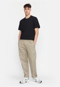 Zwarte katoenen t-shirt met korte mouwen, gecombineerd met lichtbeige broeken. Model draagt zilveren en groene sneakers. Effen achtergrond.