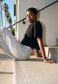 Fiatal személy fekete pólóban és világos farmerben ül kinti lépcsőn, a háttérben pálmafák, a kamerába néz.
