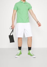 Grön polo shirt, vita shorts, gröna högskor, håller en liten svart väska. Enkelt mönster, korta ärmar och avslappnad stil.