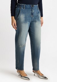 Vaqueros de denim con un lavado azul desgastado, que presentan un ajuste relajado, cinturilla elástica, bolsillos delanteros y detalles desgastados. Combinados con tacones plateados.