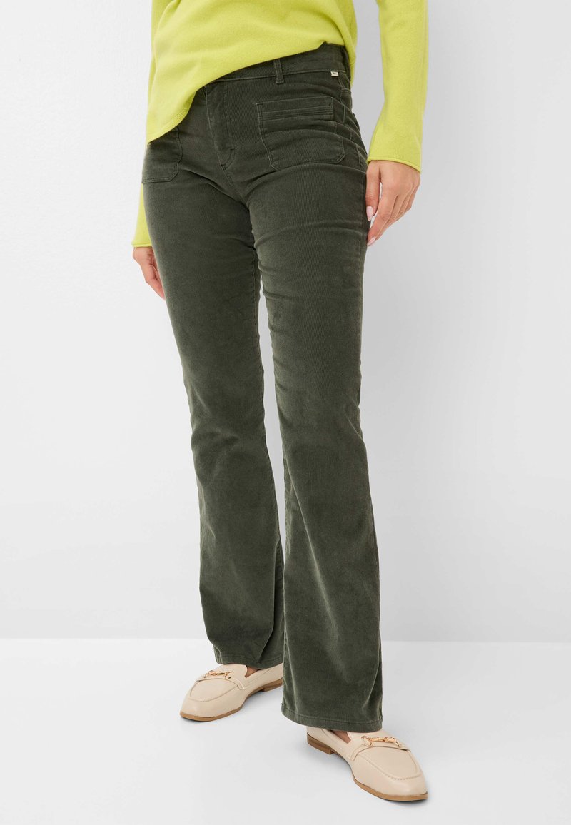 Pantalons en velours côtelé vert évasés avec poches avant, tissu à côtes texturé et taille mi-haute, associés à des chaussures de couleur claire.