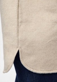 Beige, mjuk tygtopp med rundad fåll, utrustad med en sidospjäll för rörelsefrihet. Matchas med mörkt denimtyg.