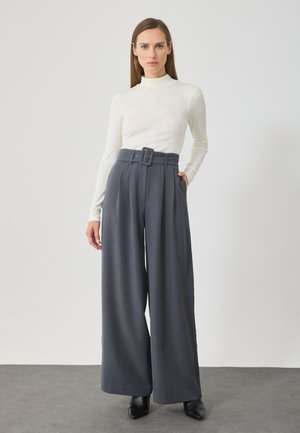 Pantaloni grigi a gamba larga con vita alta, dotati di una grande cintura, pieghe e tasche laterali. Il modello li abbina a una maglia bianca attillata.