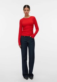 Kvinne med mørkt hår i hoop-øreringer, iført en tettsittende rød langermet topp, mørke vide jeans og spisse sorte sko med hvite prikker.