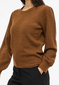 Brun strikkegenser med strukturert mønster, rund hals og lange ermer. Kombinert med sorte bukser, som viser en avslappet passform.