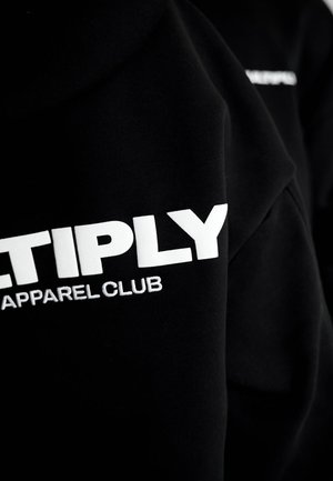 Sudadera negra con gran texto blanco que dice "MULTIPLY APPAREL CLUB", con una textura suave y elementos de diseño moderno.