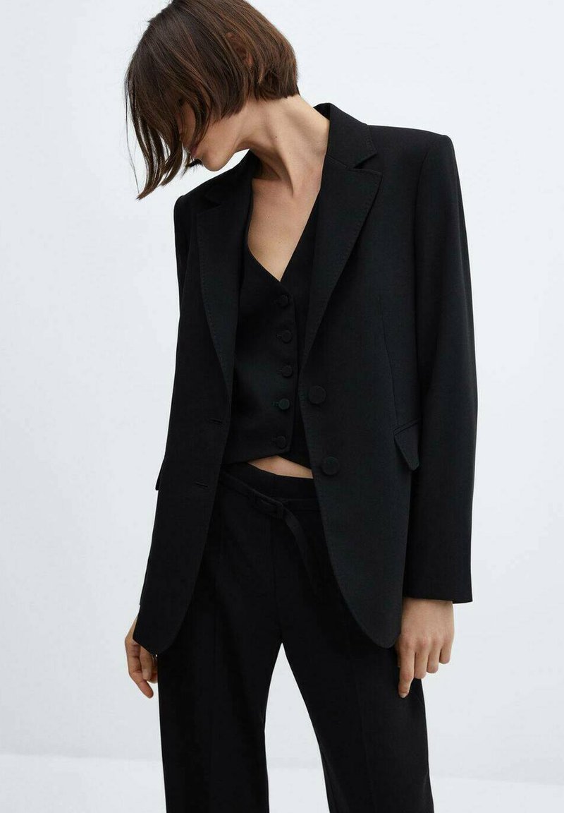 Frau in schwarzem Blazer, schwarzem knöpfbarem Top und schwarzen Hosen, die nach unten schaut, während ihr Haar teilweise ihr Gesicht verdeckt.