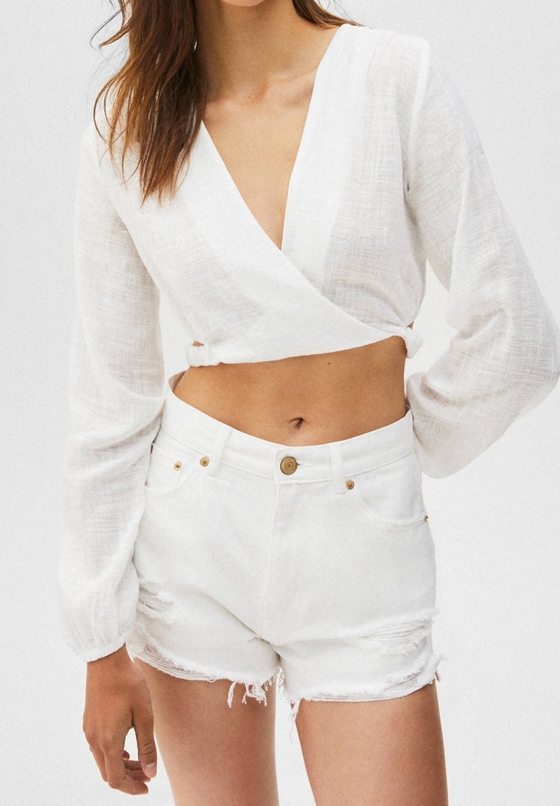 Haut court en lin blanc à manches longues et col en V, associé à un short en jean blanc déchiré avec des ourlets effilochés et des boutons dorés.