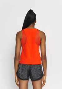 Jasnopomarańczowy tops z siateczkowymi panelami bocznymi, gładki materiał i zaokrąglony dekolt, połączony ze wzorzystymi czarnymi szortami.