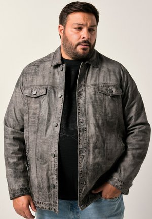 Hombre con barba que lleva una chaqueta de botón gris descolorida sobre una camiseta negra y jeans azules, de pie con una mano en el bolsillo.