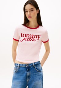 Rózsaszín pamut póló piros bordás gallérral és ujjal, piros írásos "tommy jeans" logóval. A modellen világoskék, magas derekú farmernadrág van.
