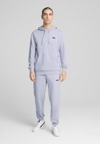 Lys lavendel hættetrøje med kengurulomme og snor, parret med matchende træningsbukser. Sort logo på brystet og venstre ben. Hvide sneakers.